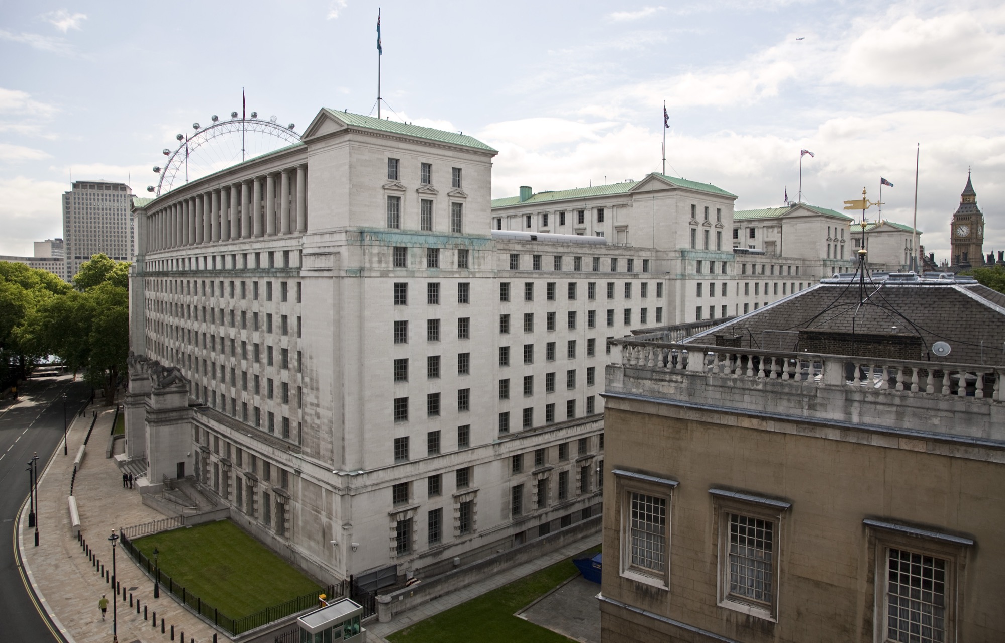 The Ministry of Defence Main Building in Whitehall, London — a vast Portland-stone block with the London Eye and Big Ben on the skyline behind it.