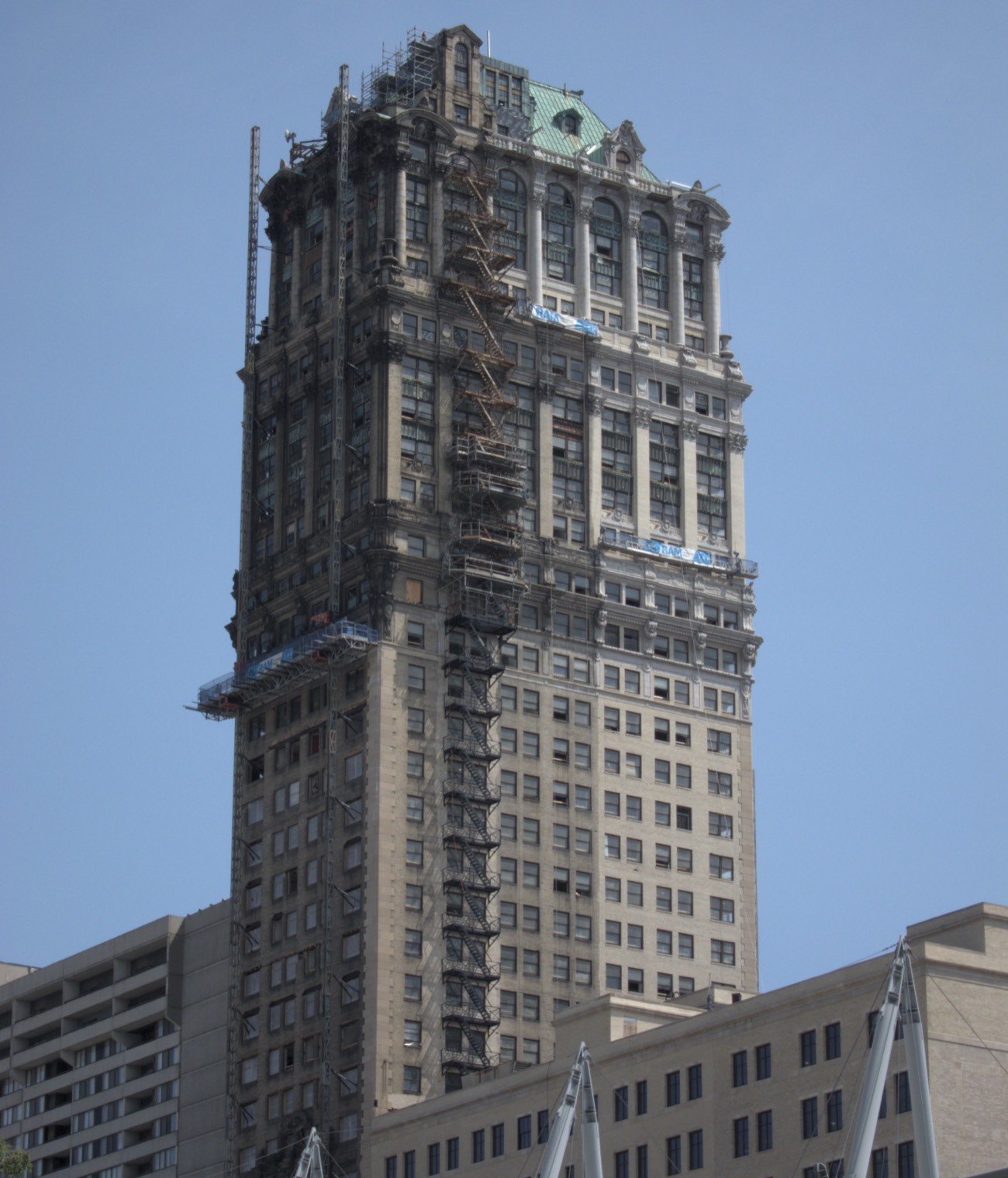 Detroit's Book Tower mid-restoration against a blue sky: a construction hoist running the full height of one flank, suspended work platforms at three levels, blackened stone on the unrestored face and a repaired green copper roof at the crown.