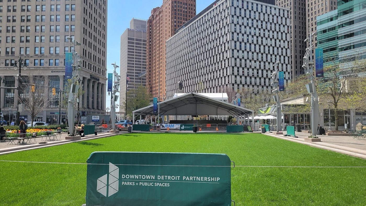 The lawn at Campus Martius Park in downtown Detroit on a spring day, ringed by office towers, with an event tent and a Downtown Detroit Partnership barrier in the foreground.