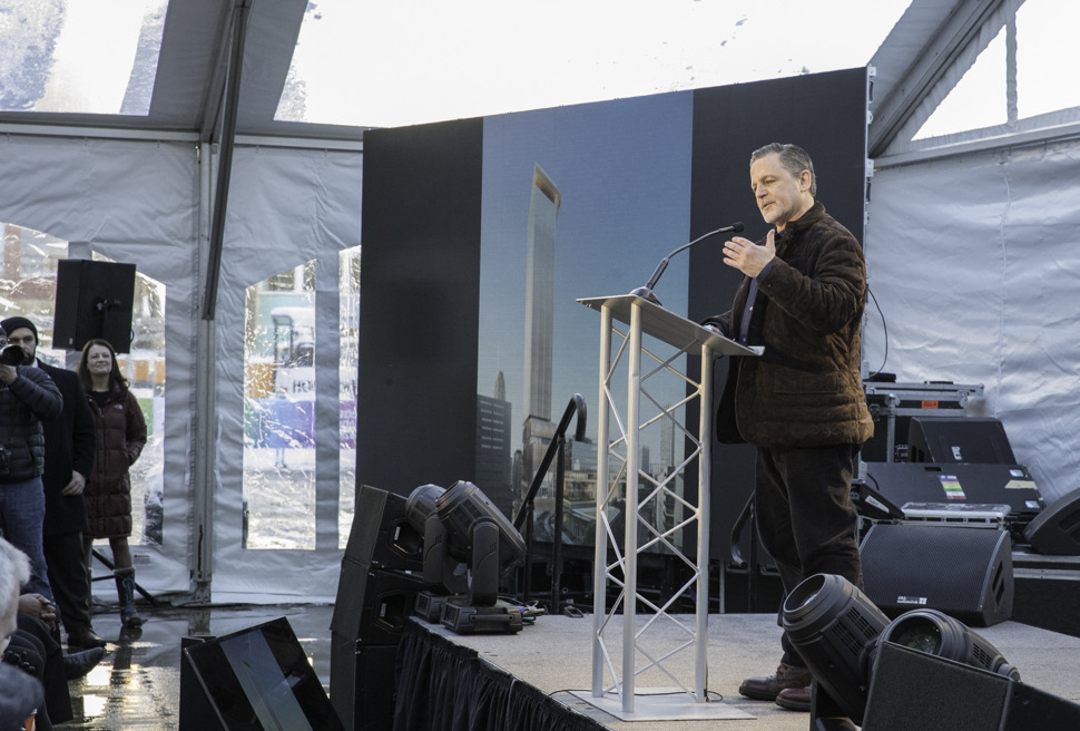 Dan Gilbert speaking at a lectern on a low stage inside a white marquee on a winter day, one hand raised, a wide screen behind him showing a rendering of a tall glass tower over a Detroit street, with photographers and guests standing at the edge of the tent.