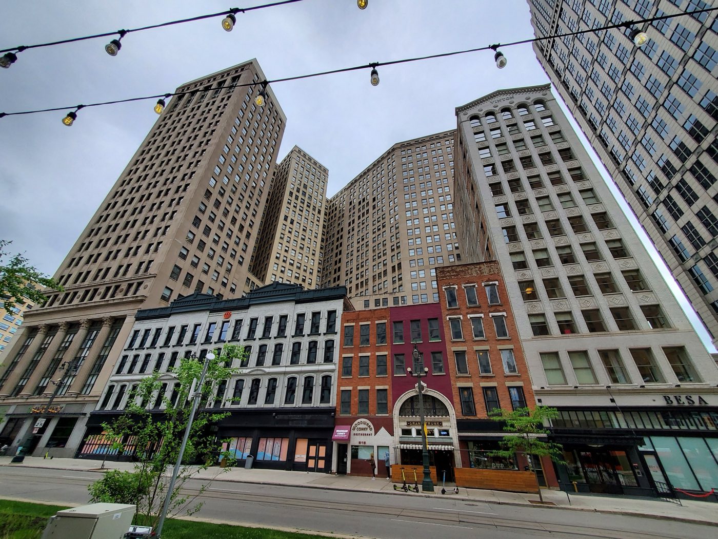 Looking up Woodward Avenue in Detroit at the limestone bulk of the First National Building, with the Vinton Building at right and a row of low nineteenth-century storefronts — a coney restaurant, a florist — along the pavement beneath them.