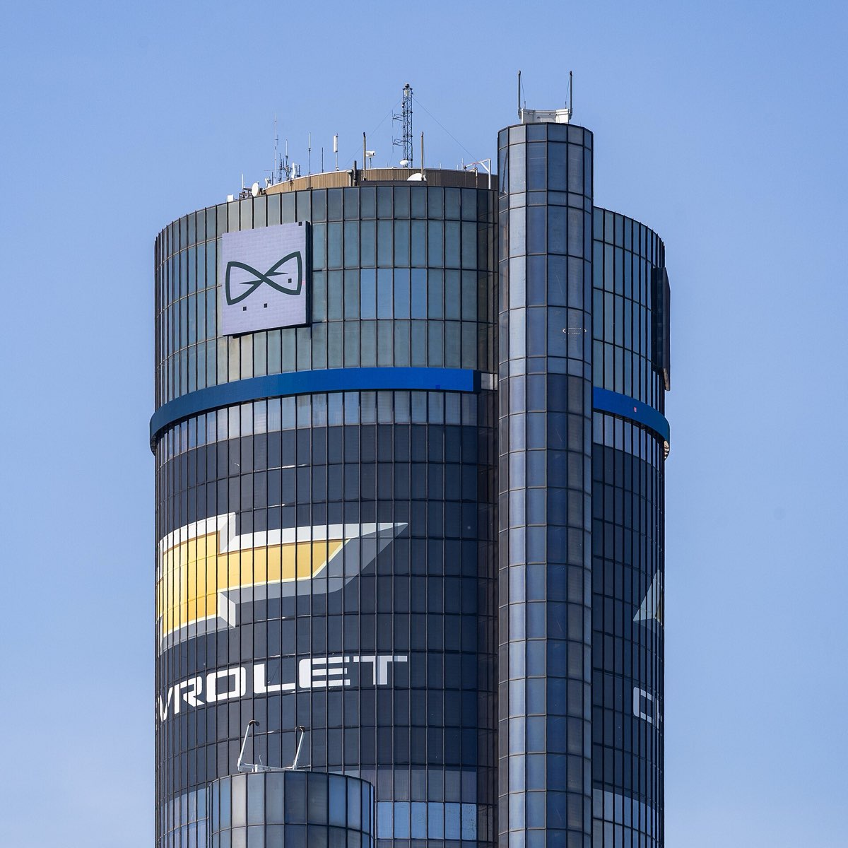 The upper floors of the Renaissance Center's central tower in Detroit against a clear blue sky, carrying a large Chevrolet emblem and a General Motors sign.