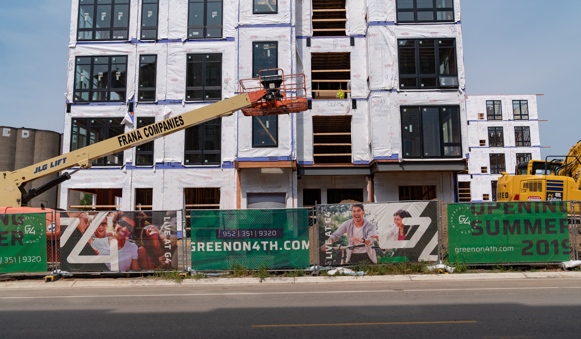 A wood-frame apartment building mid-construction in Minneapolis — Tyvek house wrap on the upper floors, windows going in, an orange boom lift raised against the facade.