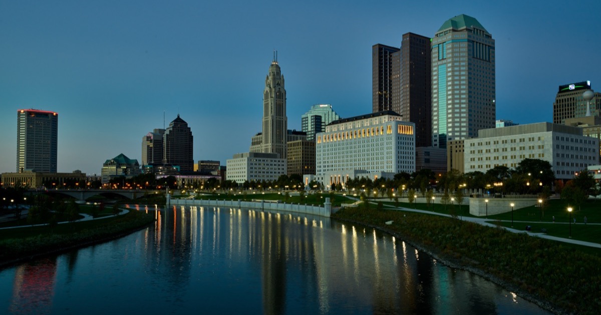 The Columbus, Ohio skyline at blue hour from the Scioto River — the LeVeque Tower and downtown office towers reflected in the water, riverfront lamps lit.
