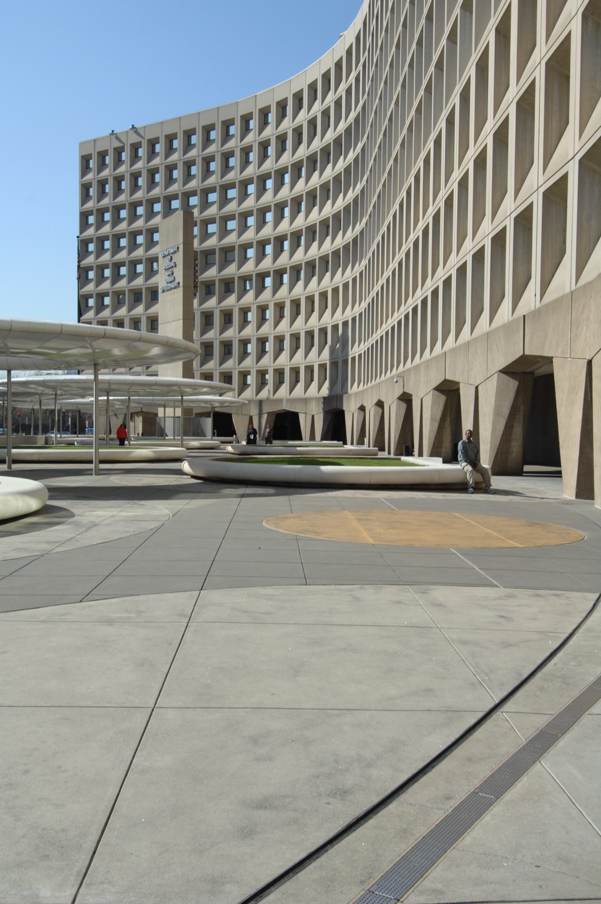 The Robert C. Weaver Federal Building in Washington — HUD’s curved, deeply coffered brutalist headquarters by Marcel Breuer, one person seated on a concrete planter for scale.
