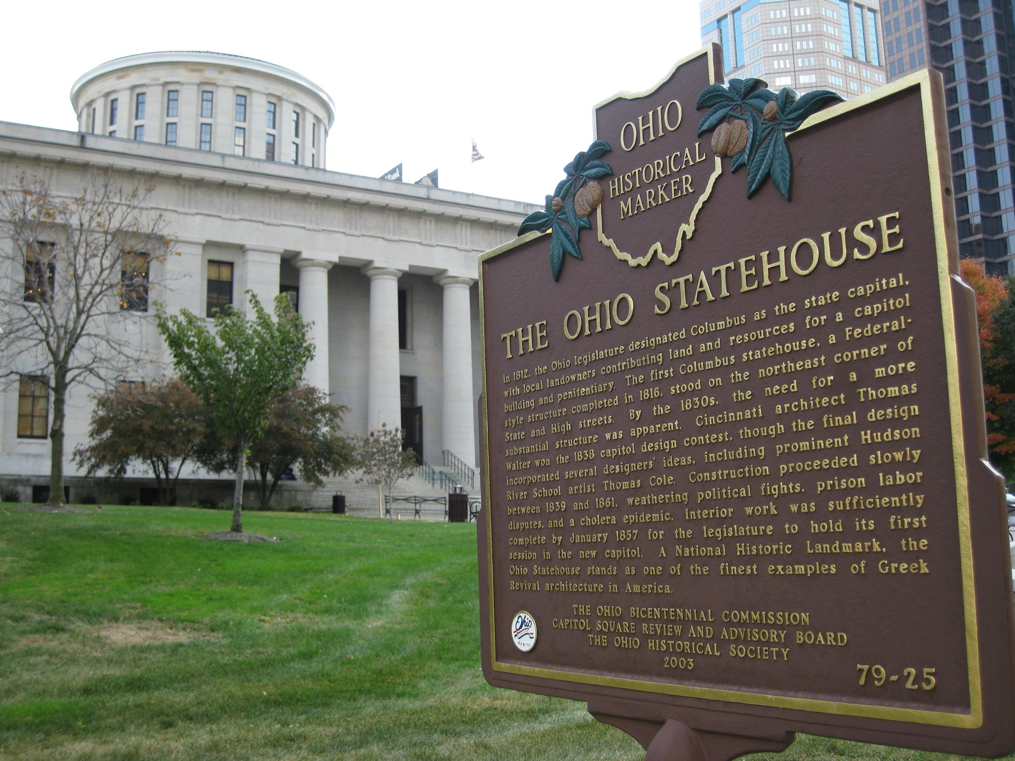 The Ohio Statehouse in Columbus behind its bronze historical marker, the Greek Revival colonnade and low dome visible past autumn trees.