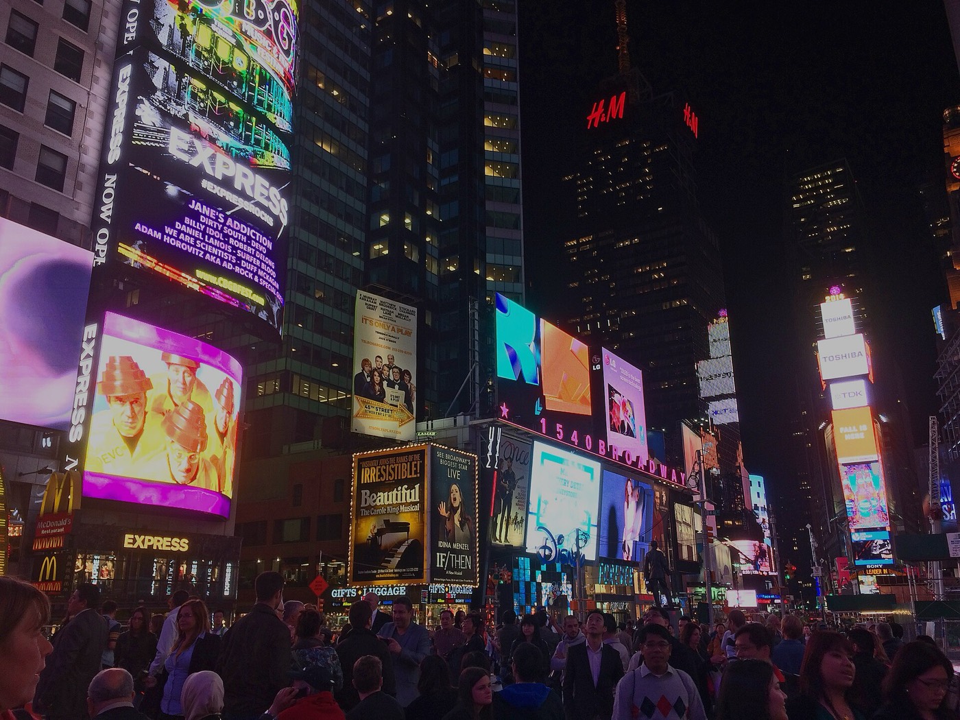 Times Square at night: a dense crowd under a wall of illuminated screens and billboards, with the words 1540 BROADWAY lit in white across the middle of the frame.