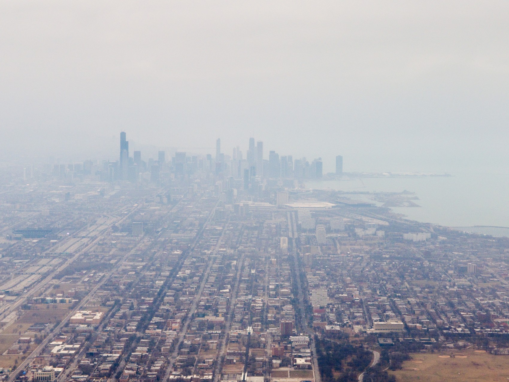 An aerial view of downtown Chicago and the Lake Michigan shoreline through haze, the towers of the Loop clustered at the water with a dense low street grid running toward the camera.
