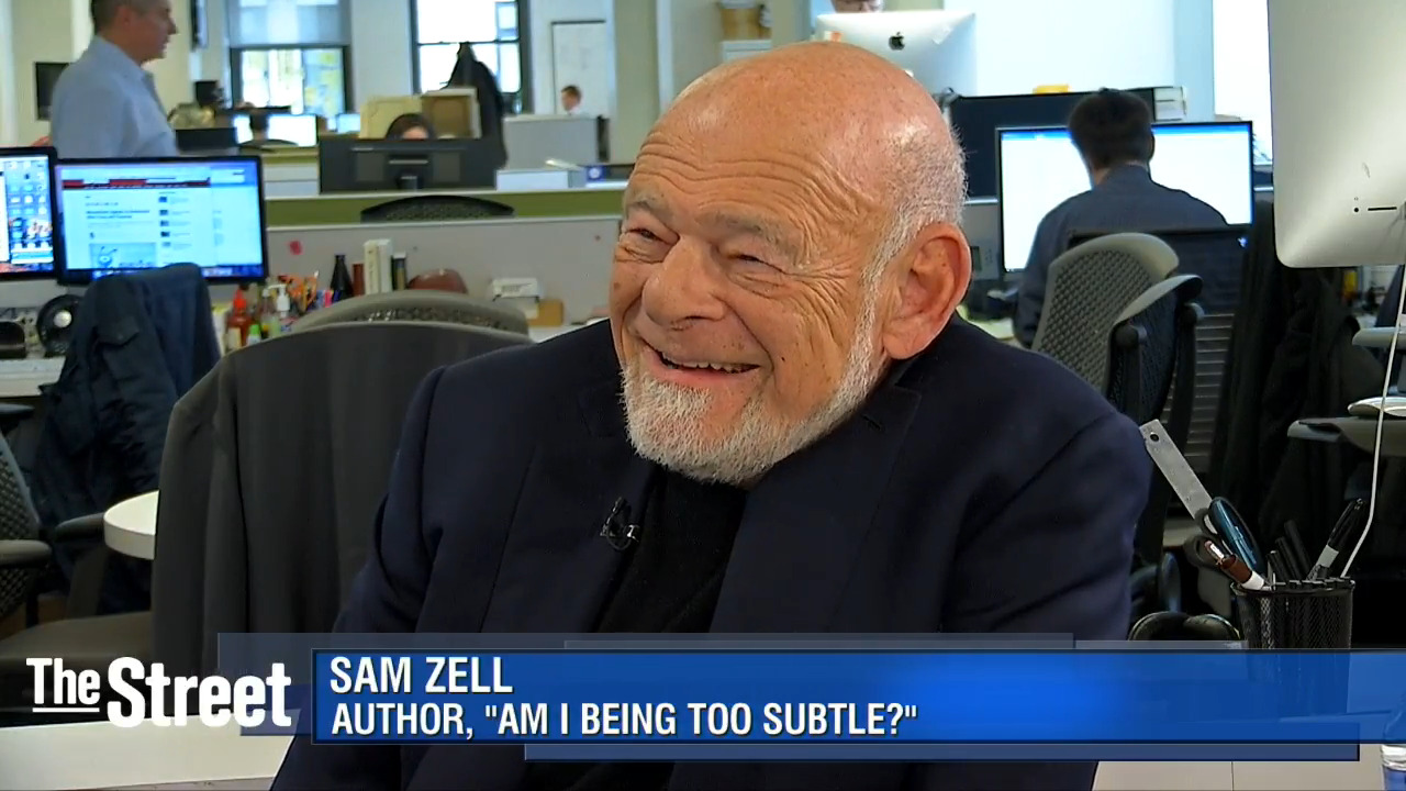 Sam Zell — bald, white-bearded, in a dark jacket over a black roll-neck — laughing during a television interview in an open-plan newsroom, with a lower-third caption naming him as the author of “Am I Being Too Subtle?”