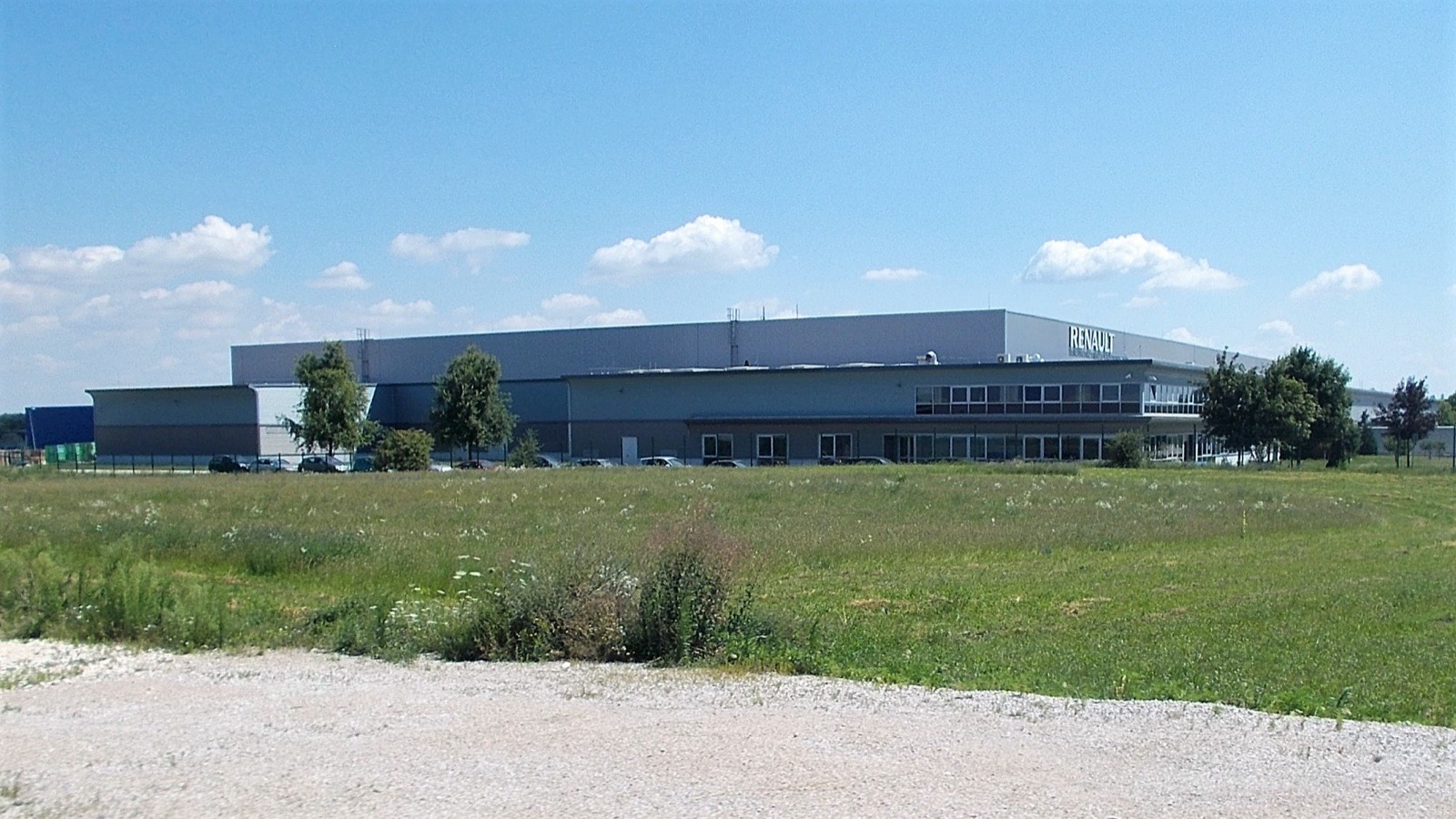 A long grey distribution warehouse with a two-storey glazed office wing and RENAULT signage, seen across a meadow under a blue sky in Győr, Hungary.
