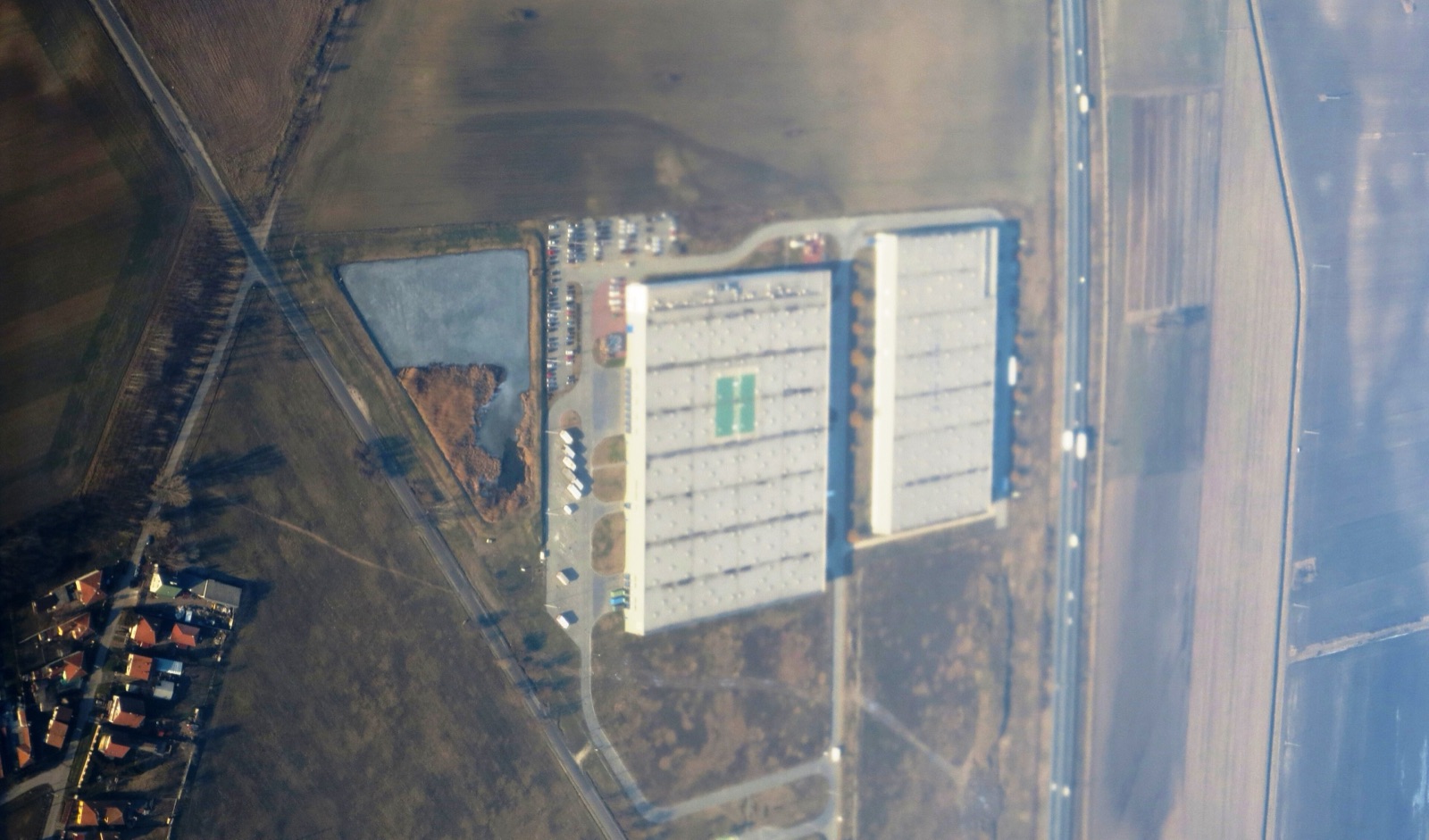 Near-vertical aerial photograph of two large white warehouse sheds and their lorry yards set in ploughed farmland, with a motorway running along one side and the red roofs of a village at the edge of the frame.