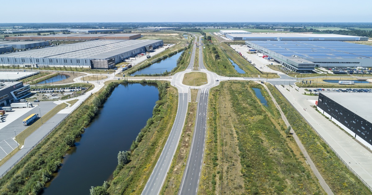 Aerial view of the Trade Port Noord logistics estate at Greenport Venlo in the Netherlands — rows of vast grey distribution warehouses, one roof covered in solar panels, lorries at loading docks, a new dual carriageway and water channels running between them.
