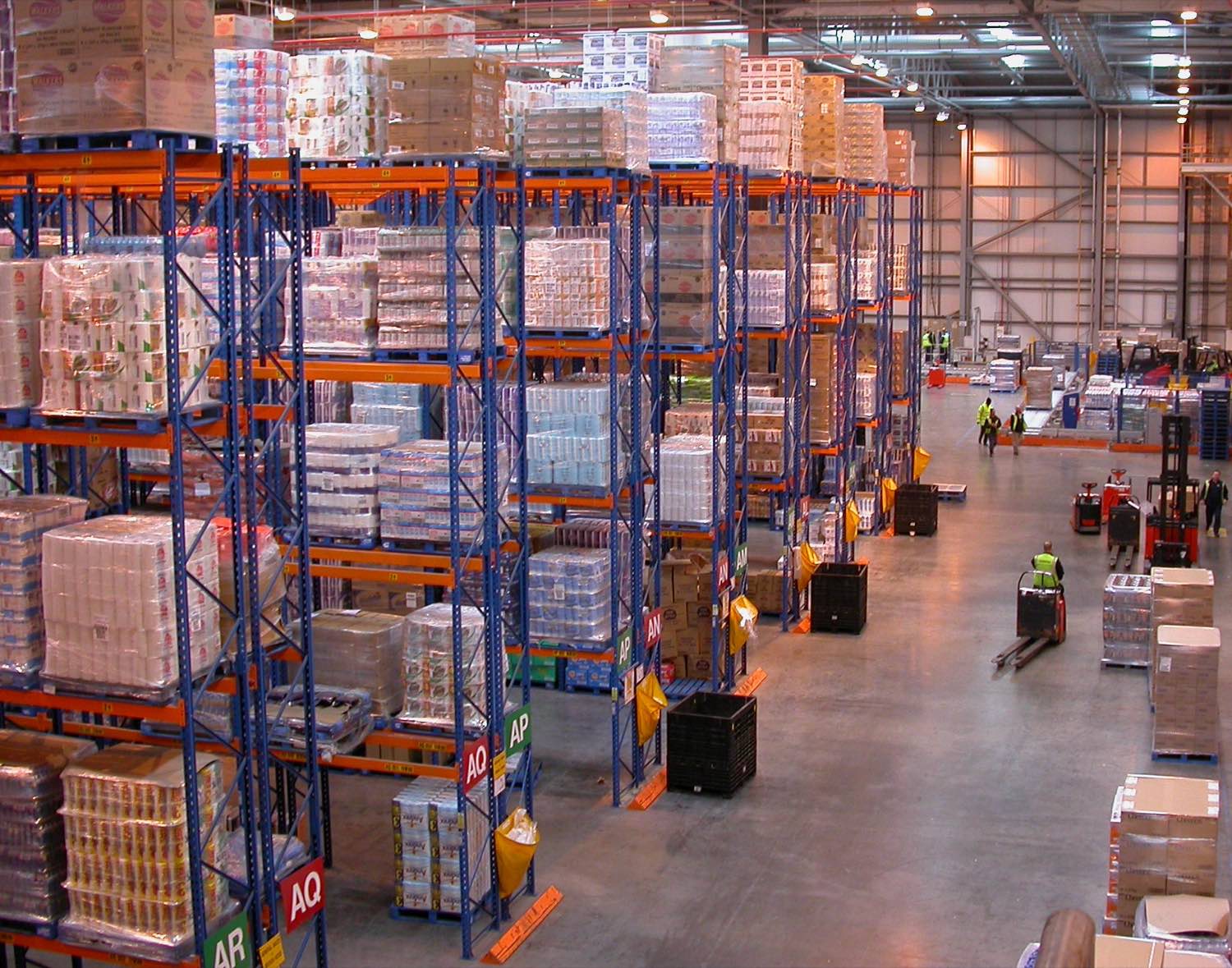 Inside a regional distribution centre — blue and orange steel racking stacked several storeys high with shrink-wrapped pallets of groceries, lettered aisle markers on the floor, and workers in high-visibility jackets moving pallet trucks down a wide concrete aisle.