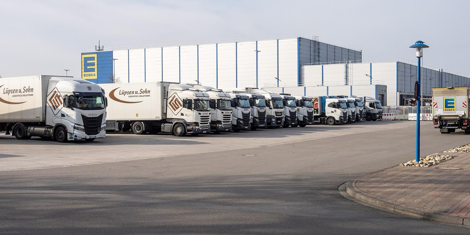 A rank of white articulated lorries and trailers parked nose-out on the apron of a large blue-and-white distribution hall carrying the EDEKA logo, under a pale winter sky in Wiefelstede, north-west Germany.