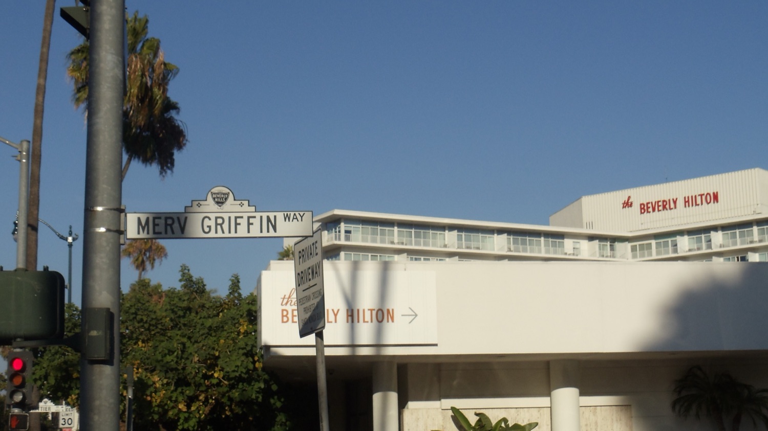 The Beverly Hilton hotel in Beverly Hills, California under a clear blue sky, its red rooftop sign reading "the Beverly Hilton", with the Merv Griffin Way street sign and palm trees in the foreground.