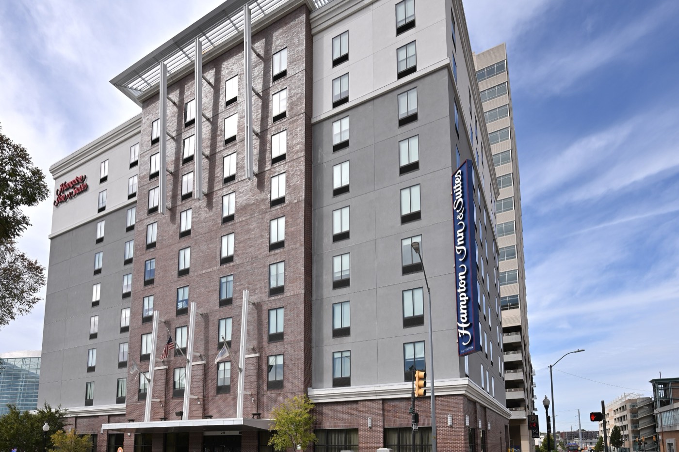 A brick and grey Hampton Inn & Suites by Hilton on a downtown Tulsa street corner under a blue sky, with the brand’s vertical blue sign on the corner of the building.