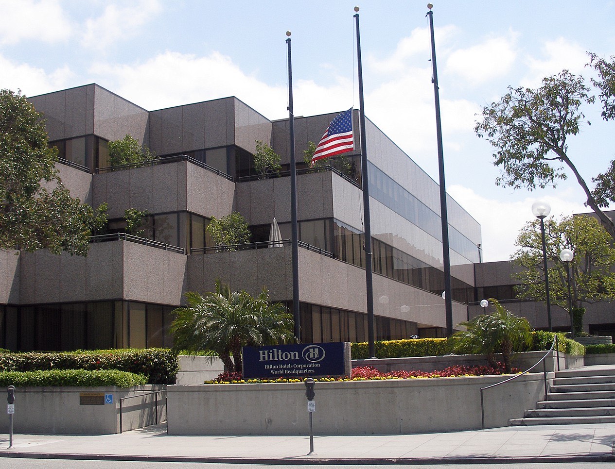 A terraced 1970s concrete office building in Beverly Hills behind palms and flowerbeds, with an American flag on a tall pole and a blue kerbside sign reading "Hilton — Hilton Hotels Corporation World Headquarters".