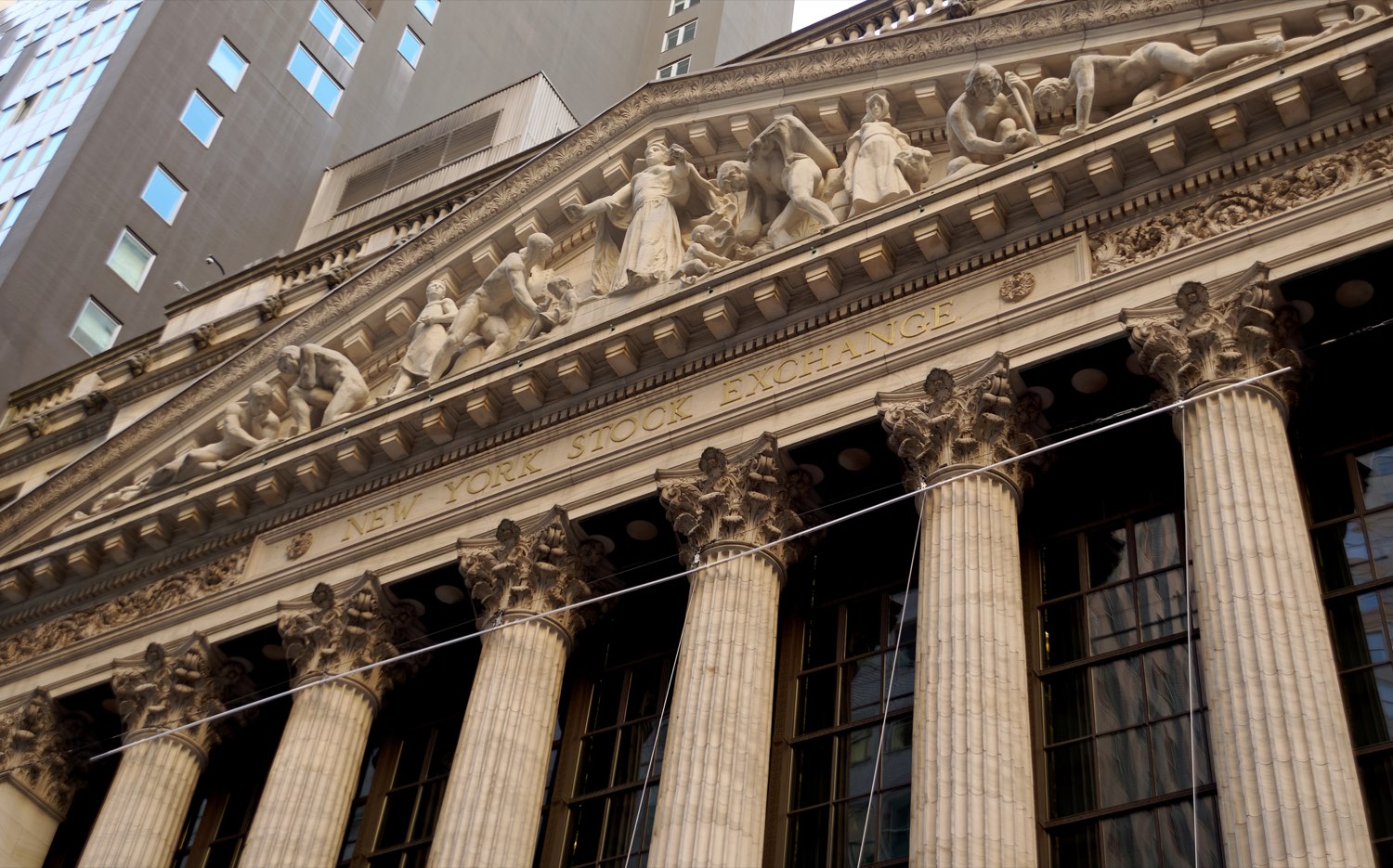 The Broad Street facade of the New York Stock Exchange from below — Corinthian columns, the carved marble pediment, and the gilded words NEW YORK STOCK EXCHANGE cut into the entablature.