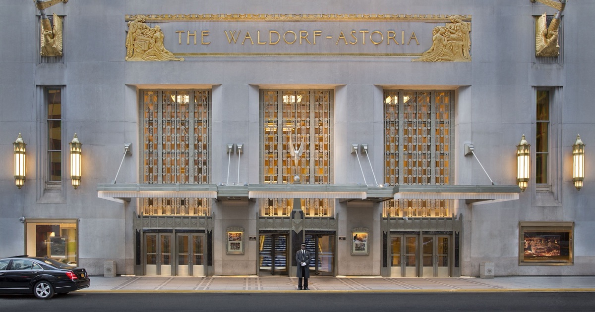 The Park Avenue entrance of the Waldorf Astoria New York at dusk — a limestone Art Deco facade with gilded relief panels reading "THE WALDORF-ASTORIA", warm light behind bronze grille doors, and a uniformed doorman standing at the centre.