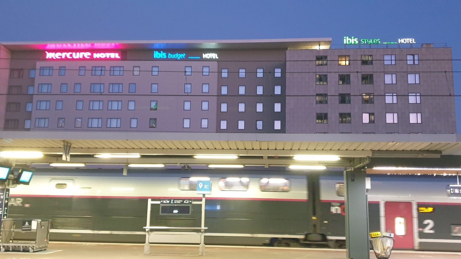 Three Accor hotel brands — Mercure, ibis budget and ibis Styles — lit in pink, blue and green along the top of one long building above a railway platform at dusk, with a TGV standing at the platform below.