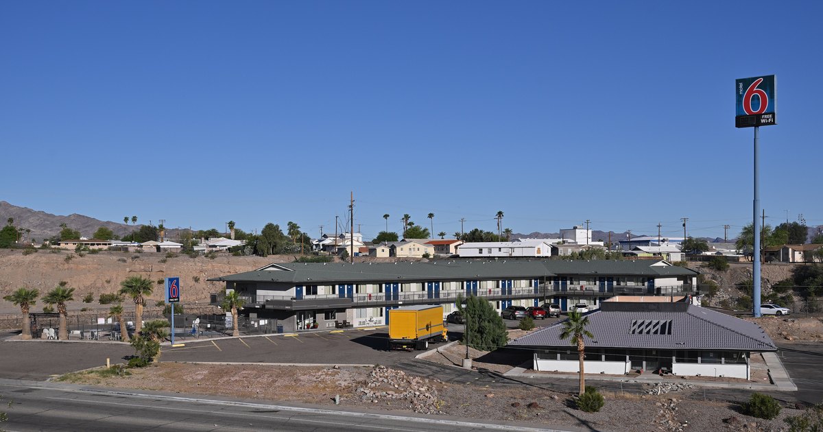 A Motel 6 property in Needles, California seen from above — a two-storey roadside motel with its tall red-and-blue 6 sign against desert hills and blue sky.