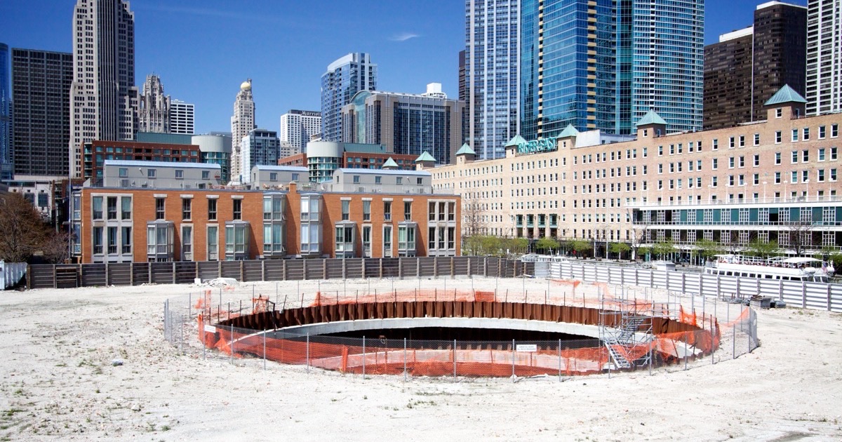 The Chicago Spire’s circular foundation excavation ringed with orange fencing on an empty gravel site, with the Streeterville skyline rising behind it.