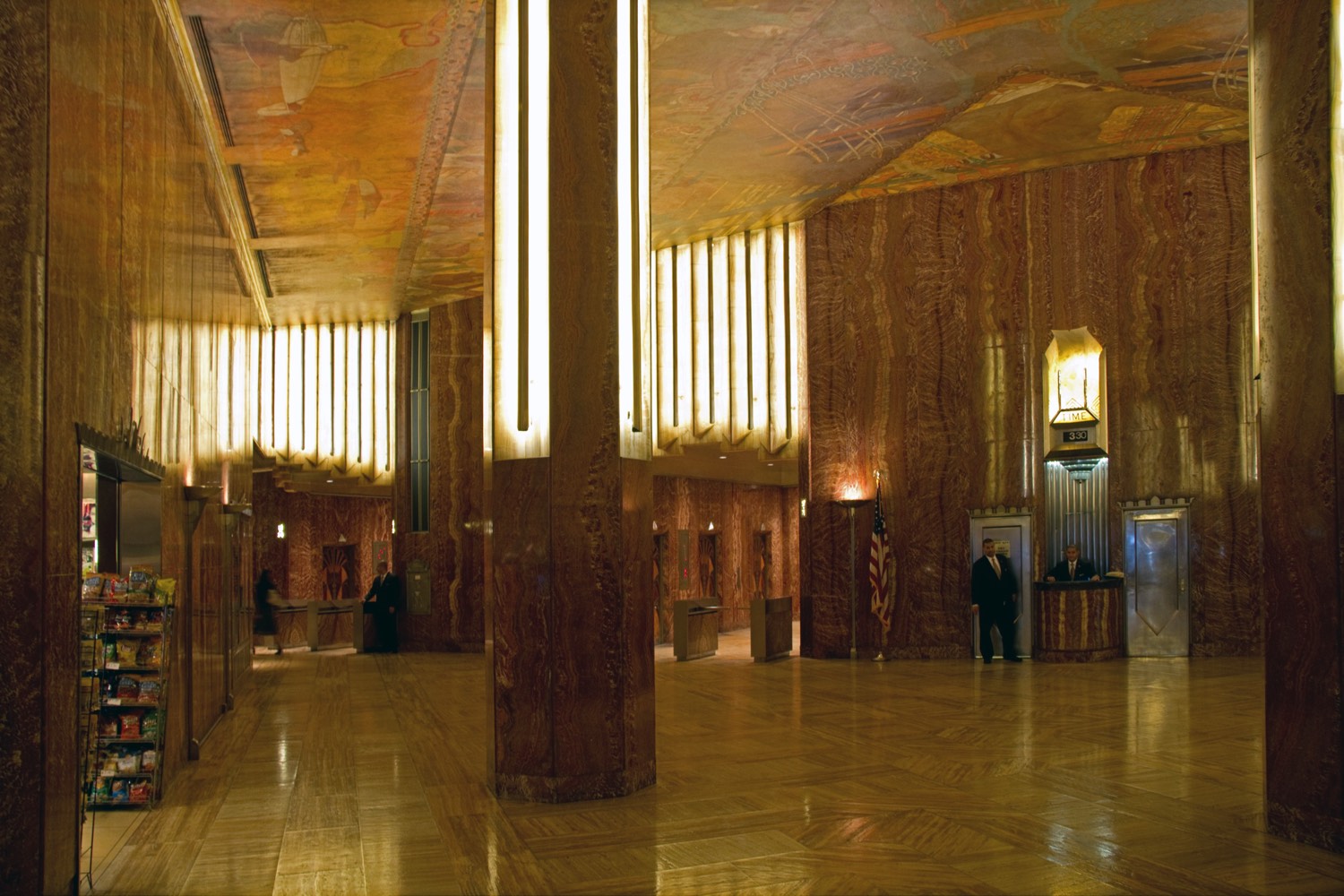 The Chrysler Building’s triangular lobby — red Moroccan marble walls, amber onyx, chrome trim and a painted ceiling mural, with visitors crossing the patterned floor.