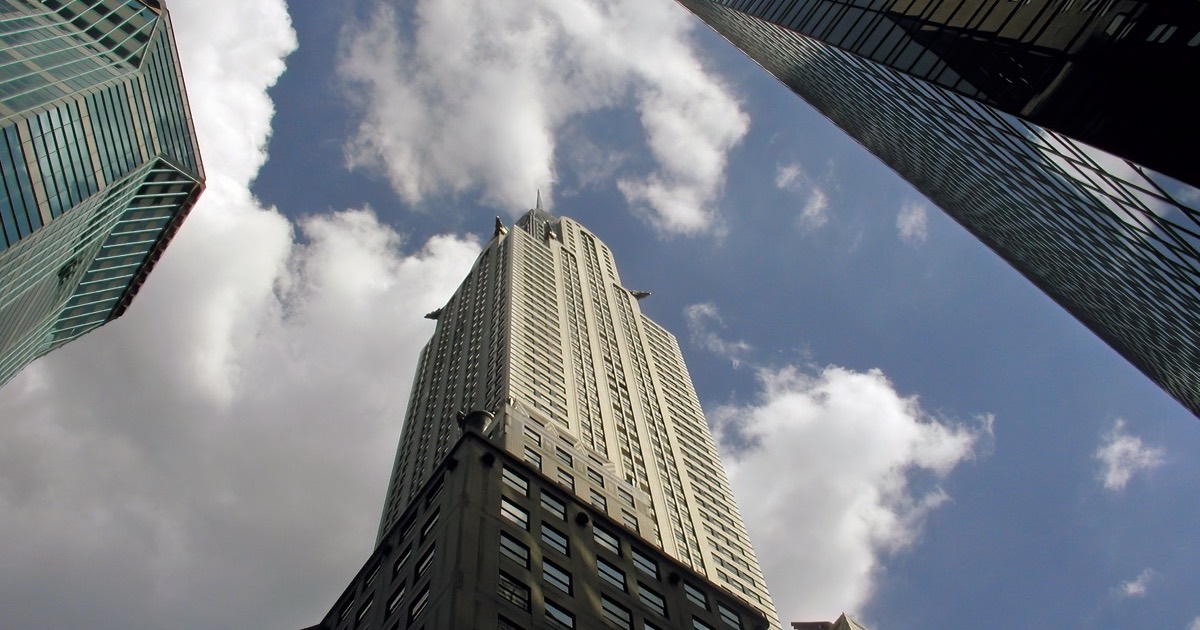 The Chrysler Building seen from street level on Lexington Avenue, its stainless steel crown and setbacks rising into a bright sky between two glass towers.