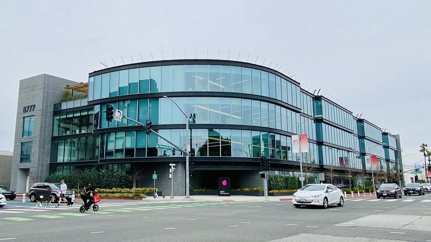 A curved four-storey glass office building at 8777 Washington Boulevard in Culver City with an illuminated Apple logo at its ground-floor entrance, seen across the intersection.