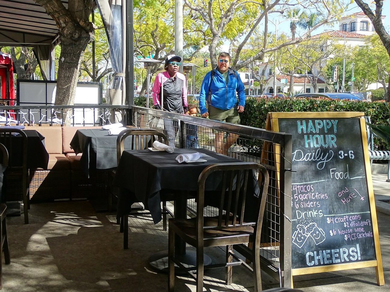 Shaded outdoor restaurant tables on a Culver Boulevard sidewalk in downtown Culver City, with a chalkboard happy-hour sign and pedestrians passing on a sunny day.