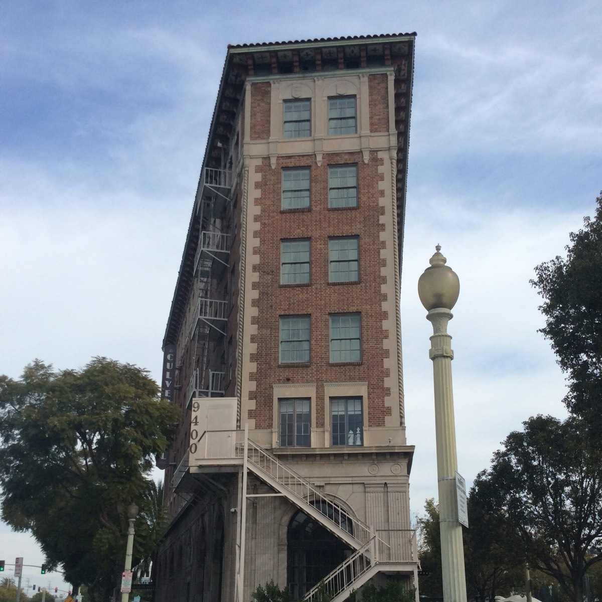 The Culver Hotel, a narrow brick flatiron building on a corner in downtown Culver City, seen from the sidewalk against a partly cloudy sky, with an exterior staircase on its front face.