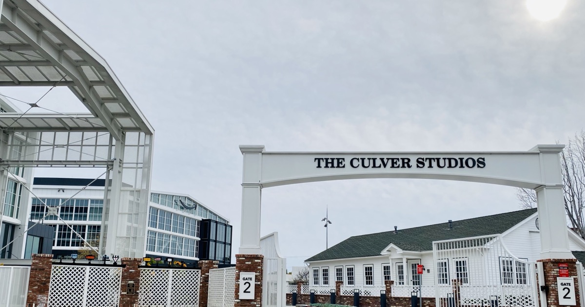 The white arched gate of The Culver Studios in downtown Culver City on an overcast day, with a glass-and-steel office building carrying Amazon's logo standing behind it to the left.