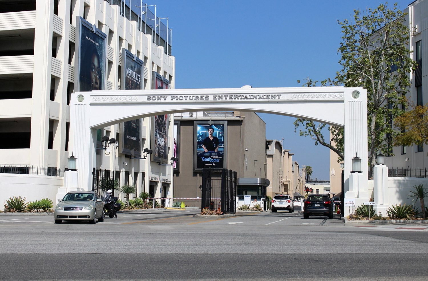 The white arched Motor Gate of Sony Pictures Studios in Culver City, lettered SONY PICTURES ENTERTAINMENT across its span, with a parking structure hung with film posters on the left and sound stages visible through the arch.
