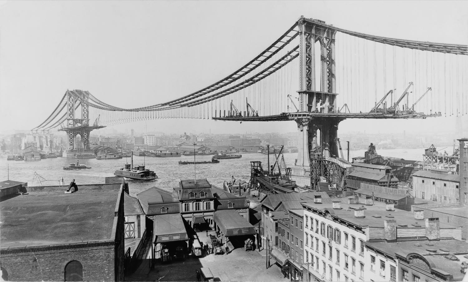 Black-and-white photograph of the Brooklyn waterfront in March 1909 — the half-built Manhattan Bridge with its deck sections still being hung from the cables, tugs and ferries on the East River, and in the foreground a ferry house marked CATHARINE FERRY beside lumber sheds and a coal company yard.