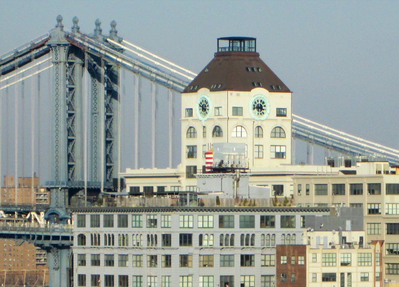 The Clock Tower building at 1 Main Street in DUMBO seen across the East River — a pale mansard-roofed tower with two visible clock faces, standing beside the stone-and-steel tower of the Manhattan Bridge above lower warehouse rooftops.