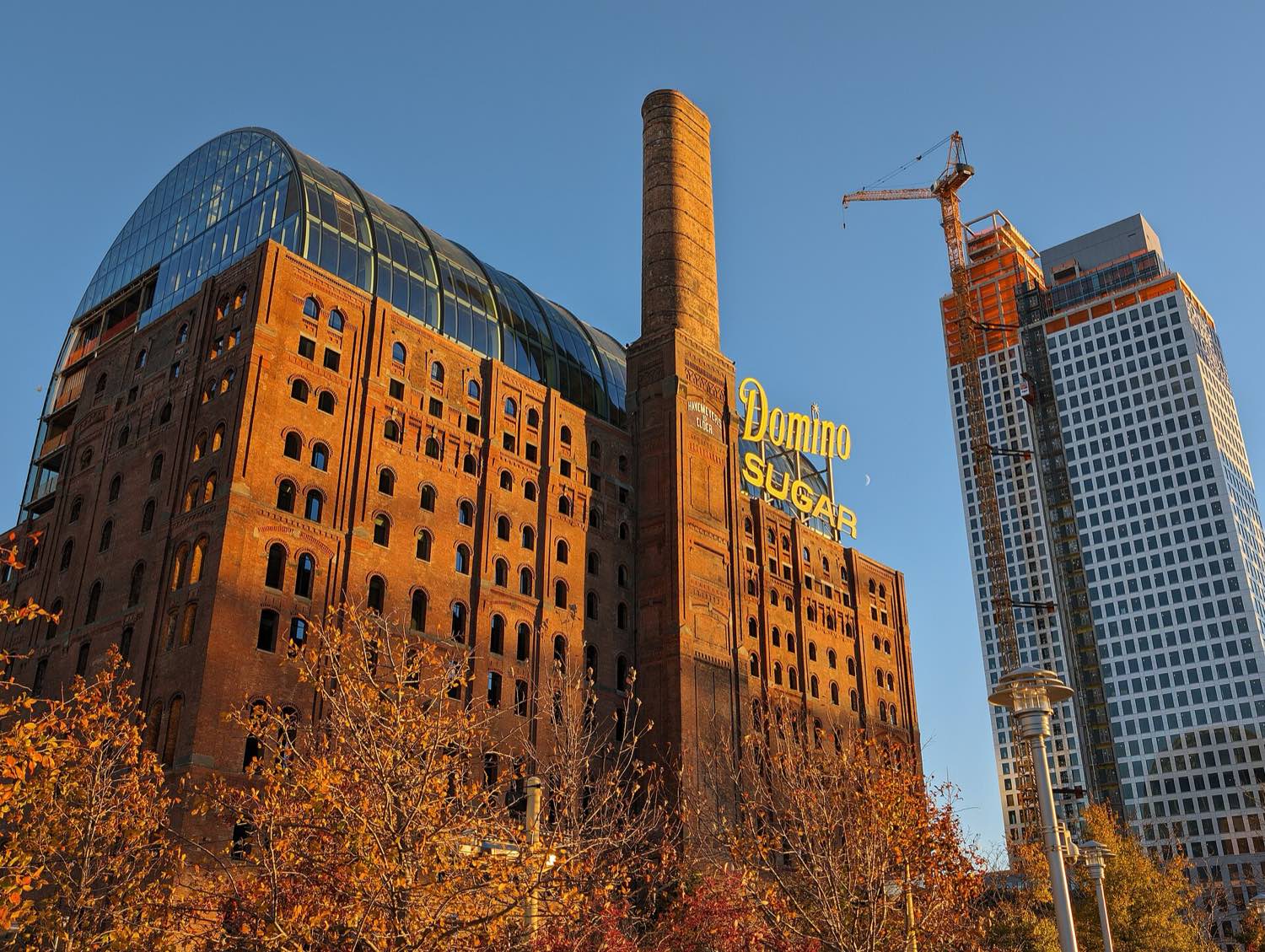 The landmarked Domino Sugar refinery in Williamsburg at sunset — a red-brick nineteenth-century mill with arched windows and a tall chimney, a curved glass office volume set inside its roofline, the yellow Domino Sugar sign on its face, and a residential tower rising under a crane beside it.