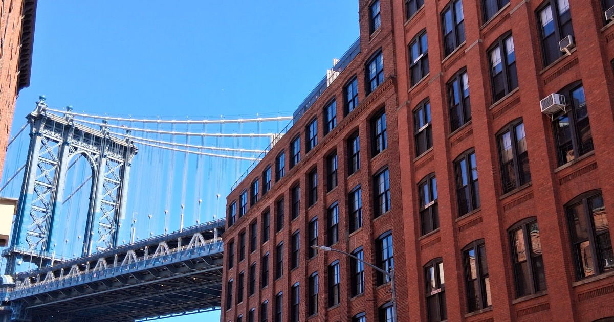 The Manhattan Bridge seen from a DUMBO street corner — its pale blue steel tower and suspension cables rising beside the tall red-brick facades of converted warehouse loft buildings, with a window air-conditioner and rows of dark sash windows, against a clear sky.