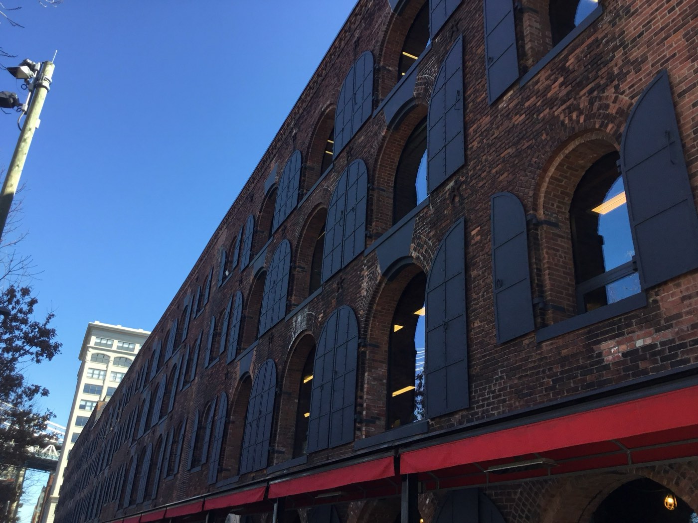 The Empire Stores on Water Street in DUMBO — a long nineteenth-century brick warehouse with rows of round-arched openings closed by dark iron shutters, restored with red awnings and lit interiors at street level.