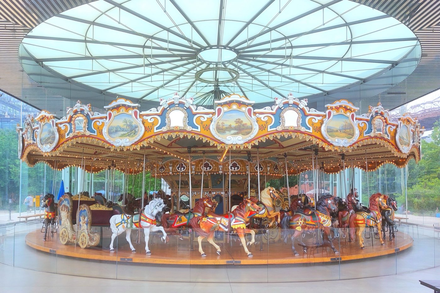 Jane's Carousel in Brooklyn Bridge Park — a restored 1922 carousel with painted horses and gilded scenery panels, standing inside a transparent glass-and-steel pavilion with a domed white canopy overhead.
