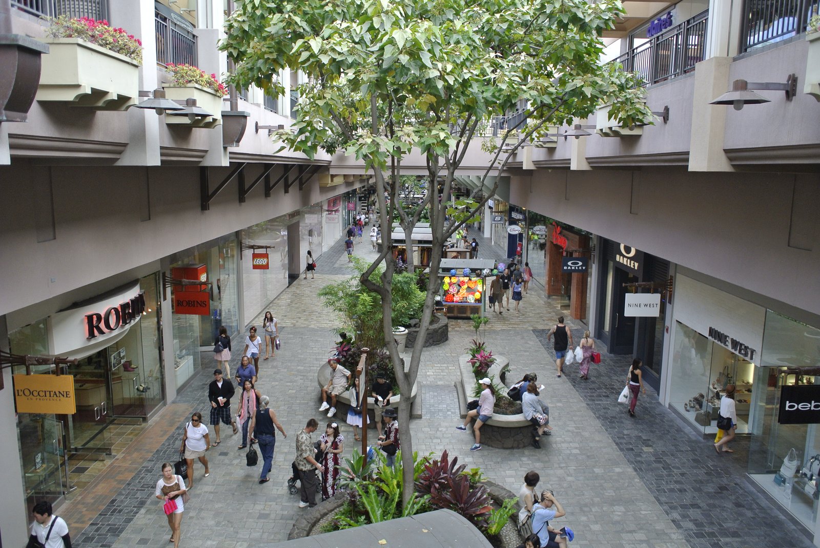 An open-air shopping promenade with trees growing in the walkway and multi-level storefronts on either side, sky visible overhead.