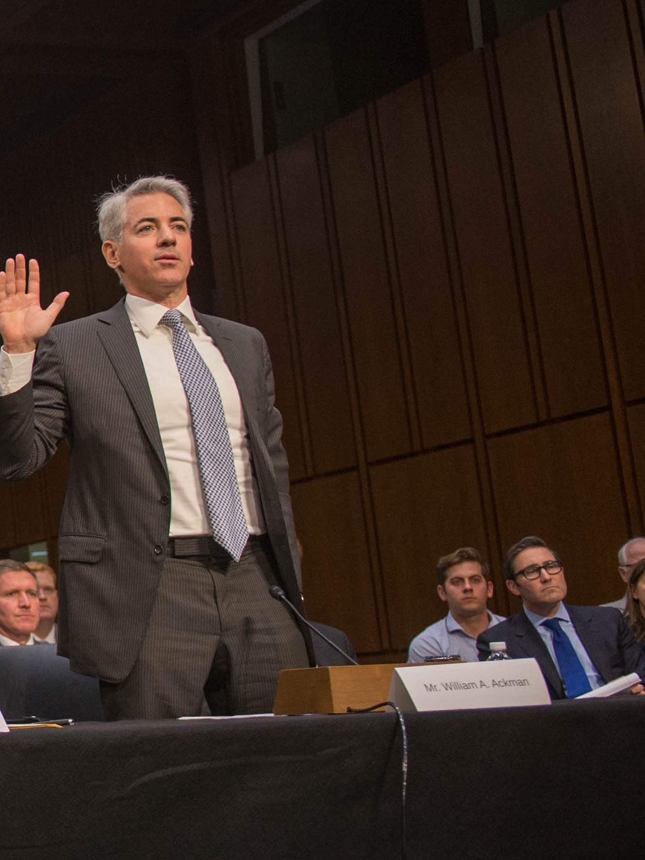 Bill Ackman stands with his right hand raised, being sworn in at a Senate hearing table, a nameplate reading “Mr. William A. Ackman” in front of him.