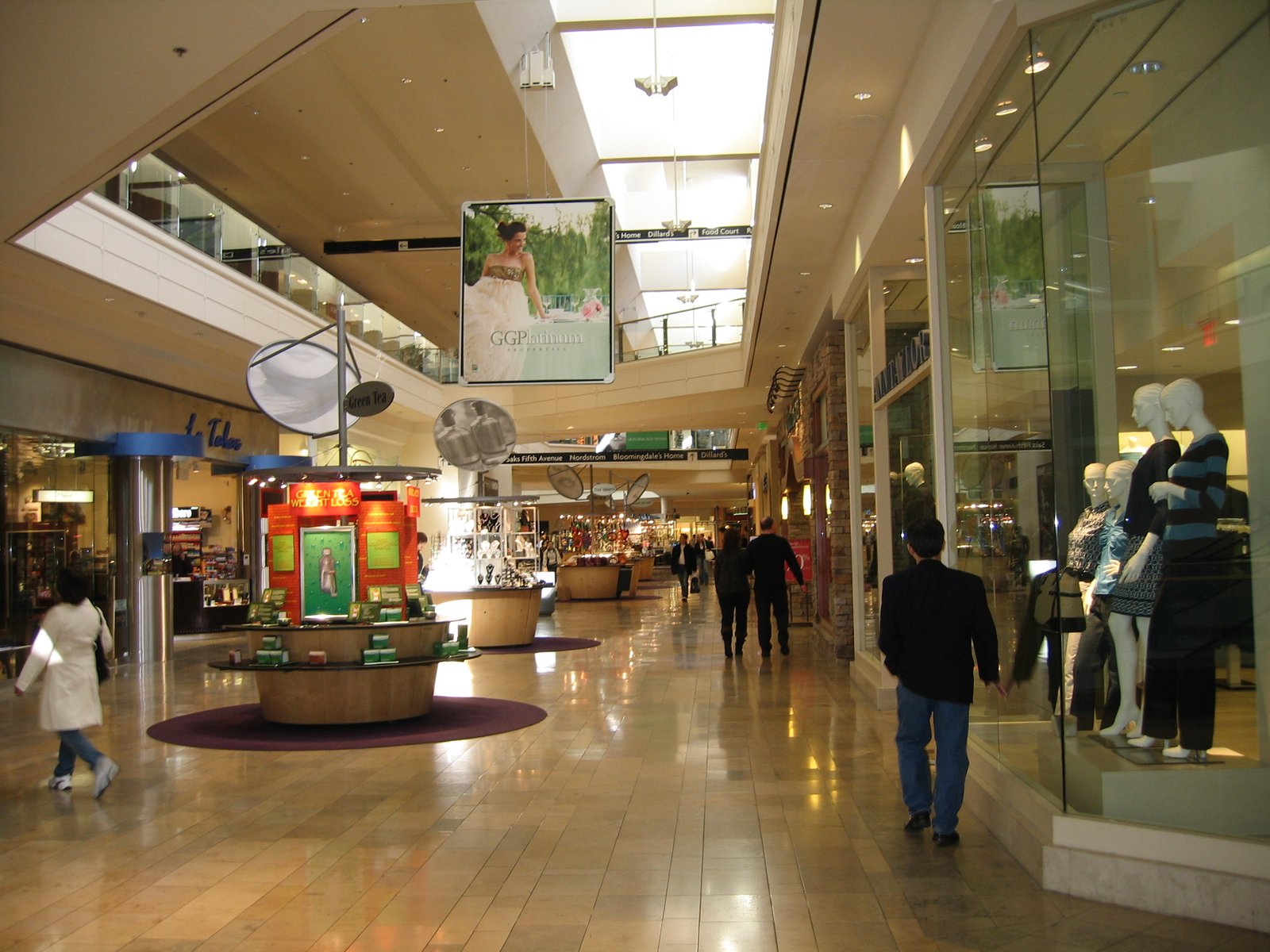 A mall corridor lined with storefronts under skylights, with a few shoppers walking through.