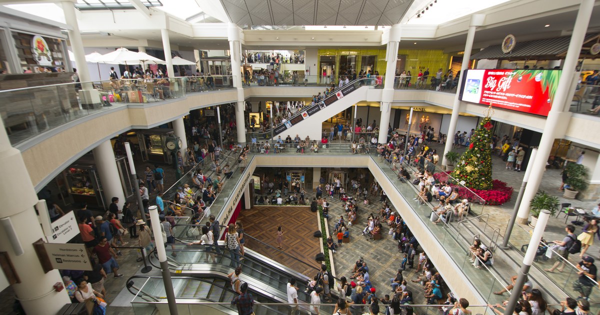 A packed, multi-level open-air shopping mall atrium with escalators, shoppers on every level, and a large decorated Christmas tree.