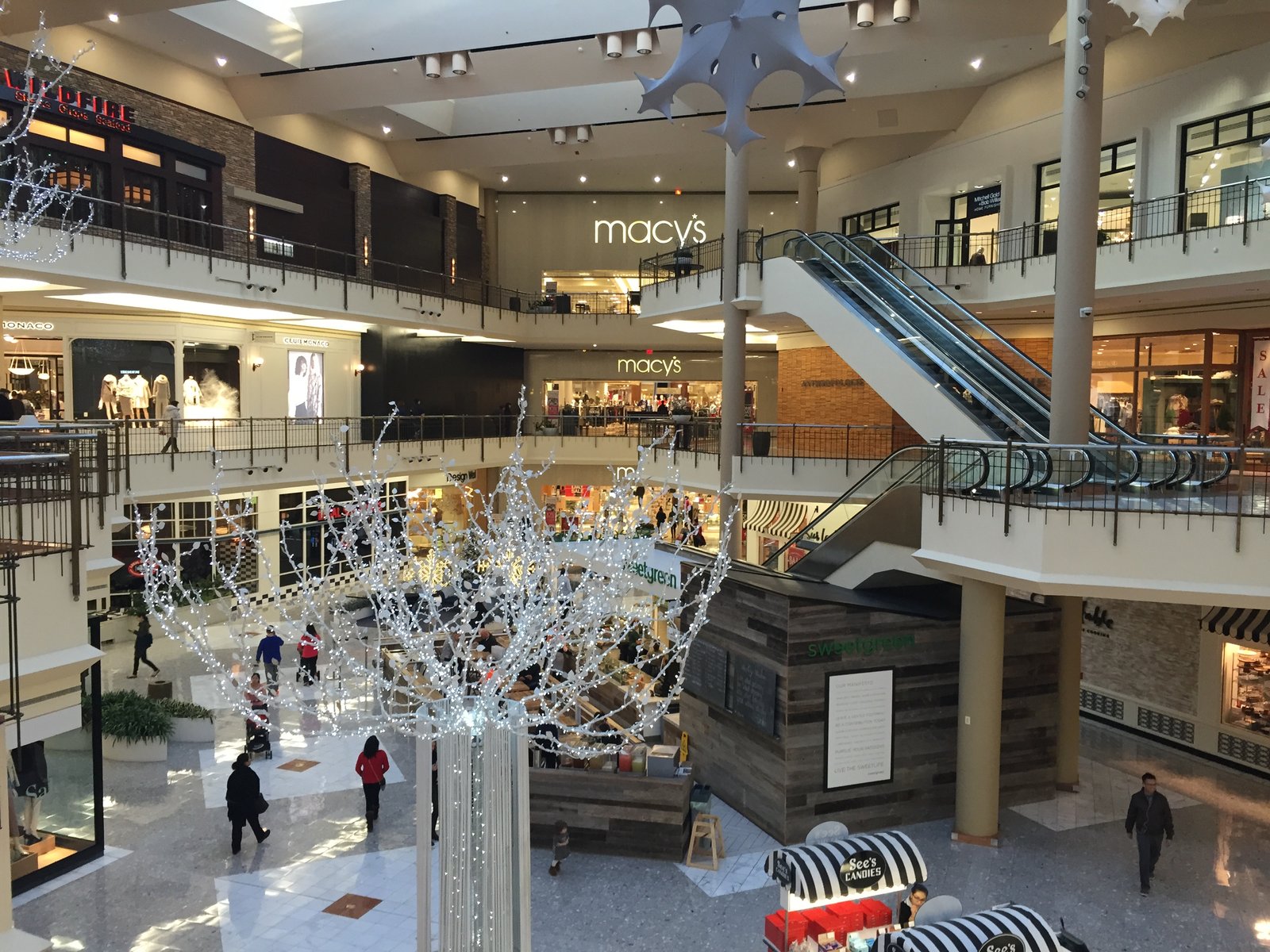 A multi-level shopping mall interior decorated for the holidays, with a Macy’s storefront sign and shoppers on the lower level.