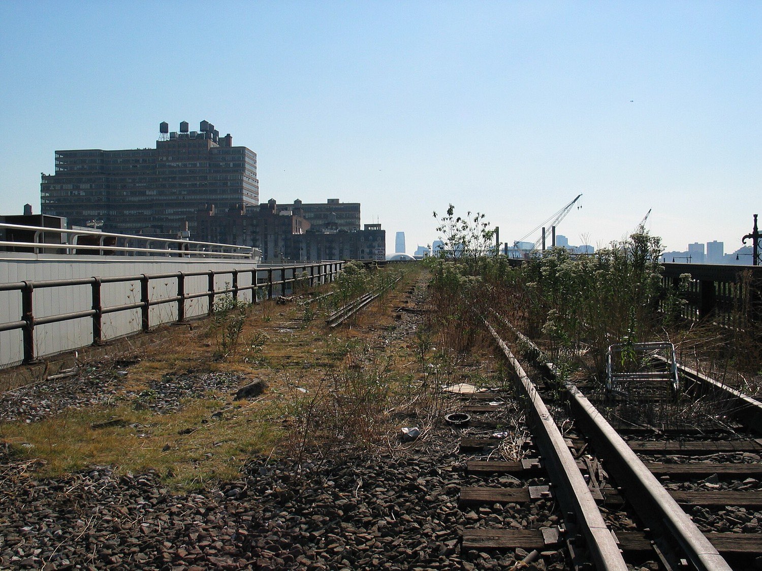 The abandoned High Line in October 2005 — rusted rails and ballast running into the distance, overgrown with dry weeds and self-seeded shrubs, an abandoned shopping trolley to one side, low West Chelsea rooftops beyond.