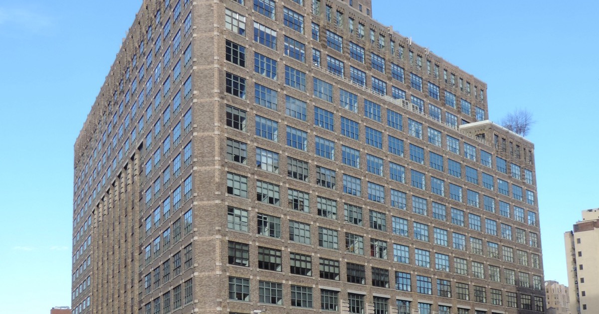 The brick massing and window grid of One Hudson Square, 75 Varick Street, against a blue sky — a printing-district block in Trinity Church’s campus.