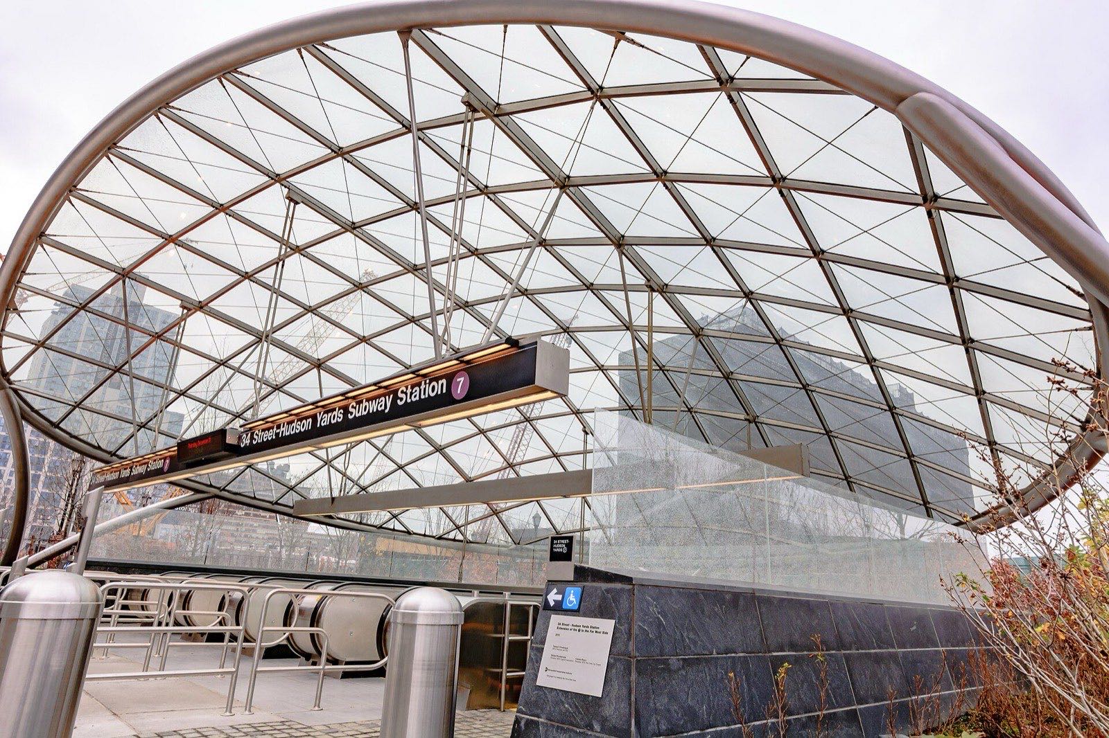 The curved glass-and-steel canopy over the 34th Street–Hudson Yards subway station entrance, with an illuminated sign reading 34 Street-Hudson Yards Subway Station 7 above the escalators.