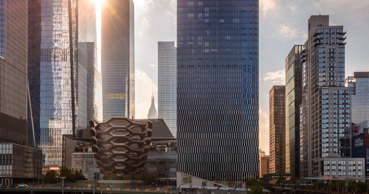 The Hudson Yards cluster of glass towers backlit by a low sun, with the Vessel’s honeycomb structure silhouetted in front and the Empire State Building’s spire visible between two towers in the distance.