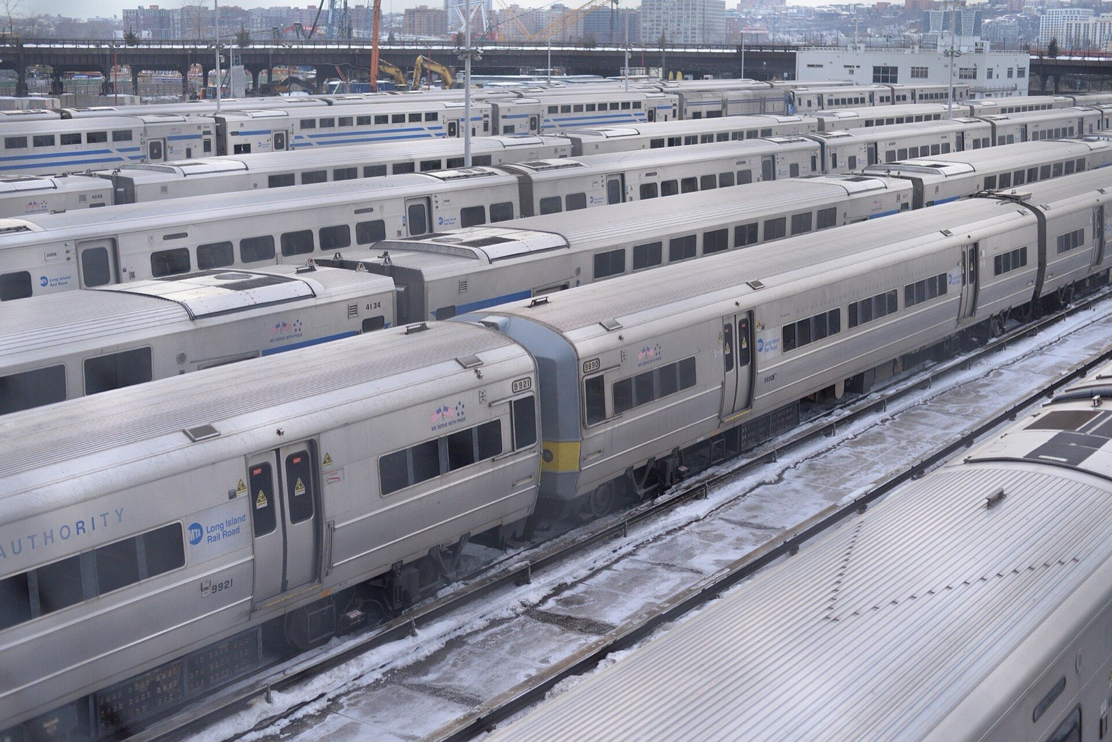 Rows of Long Island Rail Road trains parked on tracks in the open West Side Yard, snow on the ground, construction cranes and the New Jersey shoreline in the background.