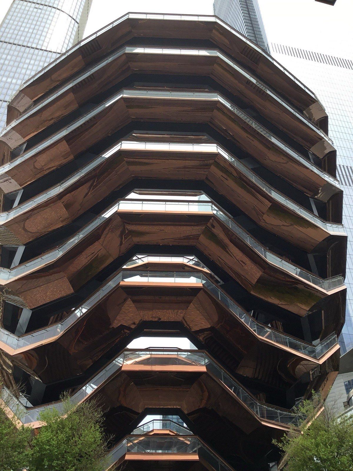 Looking straight up at the Vessel, a spiralling copper-toned honeycomb of interlocking staircases, with a glass skyscraper rising behind it.